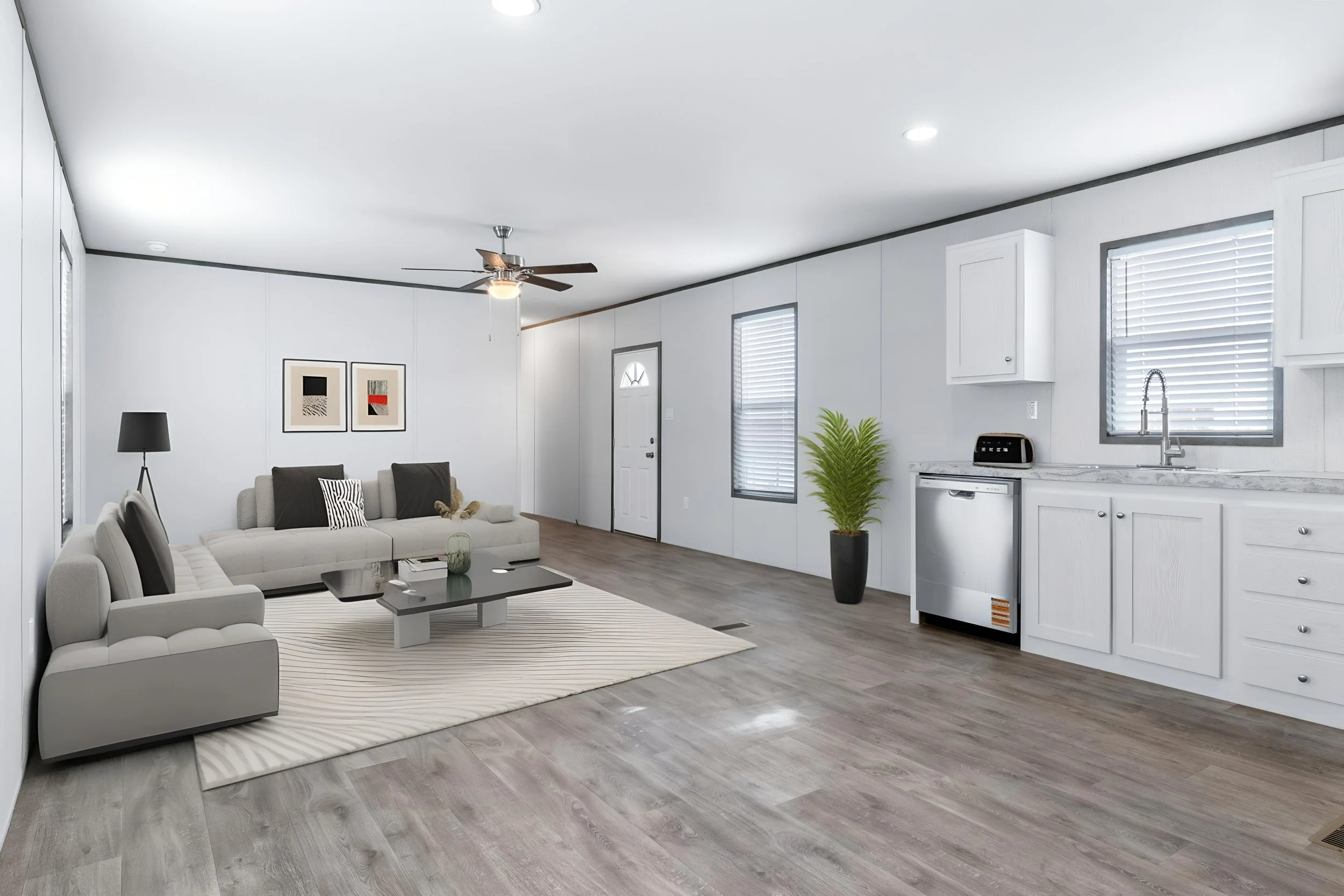 Open-concept living room and kitchen with gray wood flooring, white walls, a gray sectional sofa, a black coffee table, a white area rug, framed artwork, a floor lamp, and a vaulted ceiling with a ceiling fan. The kitchen has white cabinetry, a marbl