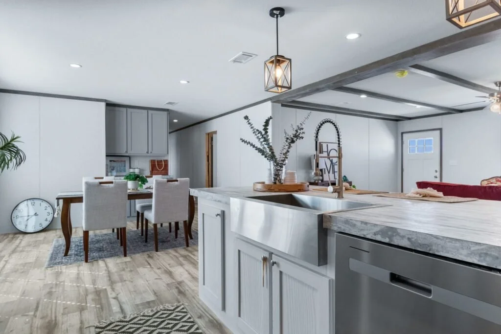 Open-concept kitchen and dining area with white cabinetry, wooden flooring, a large farmhouse sink, and a dining table with six chairs, decorated with plants and a wall clock.