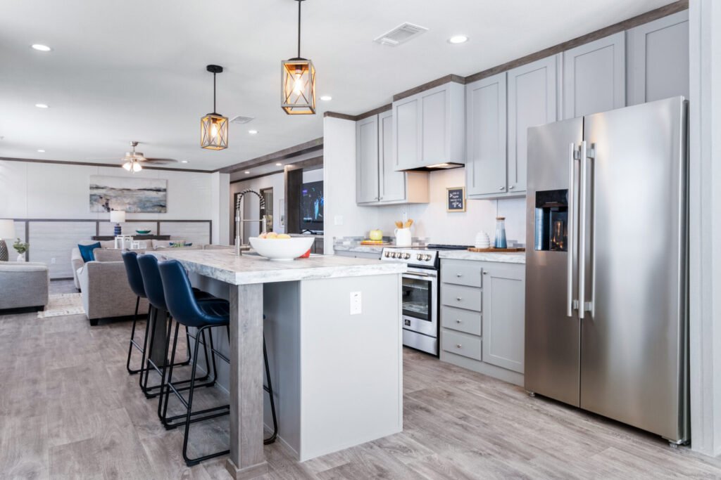 Modern kitchen with light gray cabinets, stainless steel refrigerator, white marble island with three blue chairs, pendant lights, and connected living area with a sofa and wall art.