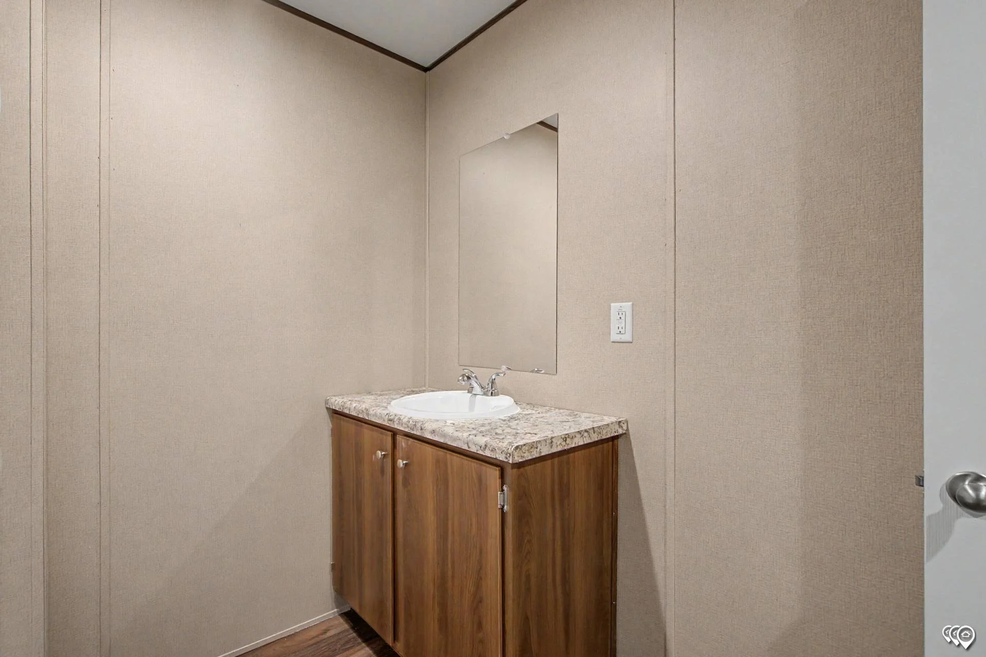 A bathroom vanity with a white sink, silver faucet, wooden cabinet, and a granite countertop, mounted on a wall with beige textured wallpaper and a mirror above.