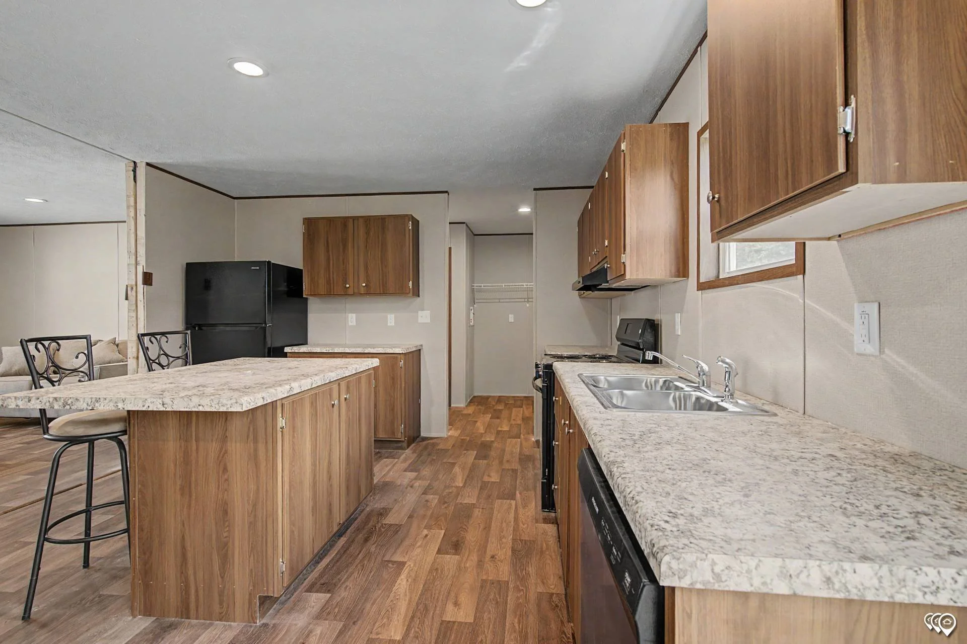 Interior of a kitchen with wooden cabinets, granite countertops, stainless steel sink, black refrigerator, and a ceiling with recessed lighting; view includes part of a dining area with chairs and a sofa.