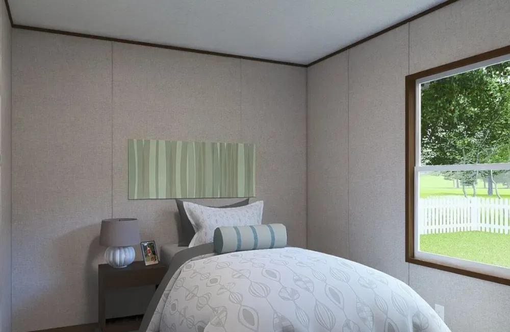 A bedroom with beige walls, a bed with white and gray bedding, a bedside table with a gray lamp and picture frame, and a window showing a grassy yard with trees and a white picket fence.