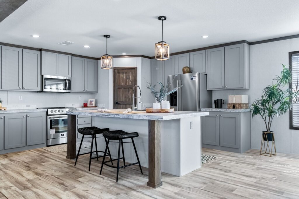 Modern kitchen with light gray cabinets, a marble island with two black stools, wooden flooring, and stainless steel appliances, including a refrigerator and microwave. Two pendant lights hang over the island, and a plant in a black and gold pot sits
