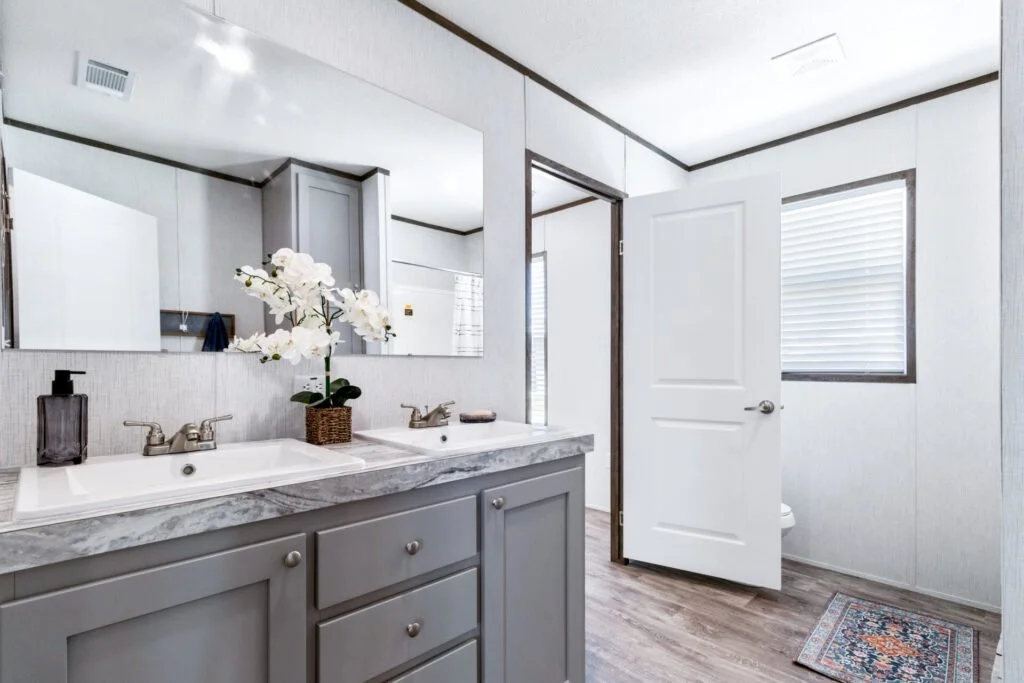 A modern bathroom with a double sink vanity, a large mirror, and a flower arrangement on the countertop. There is a window with blinds, a door partially open revealing a toilet, and a decorative rug on the wooden floor.