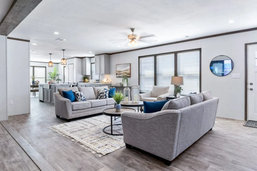 Bright living room with two gray couches, white and blue throw pillows, a round coffee table with decorations, a white armchair, a table lamp, large windows with blinds, and a view into the kitchen and dining area.