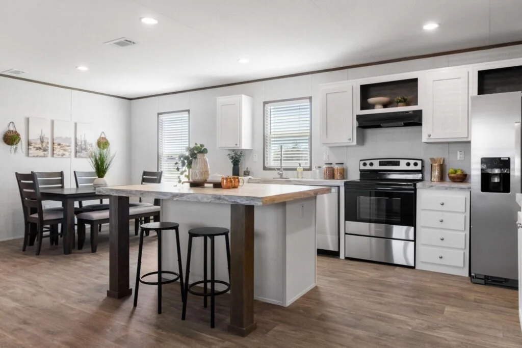 A bright, modern kitchen with white cabinets, stainless steel appliances, a marble countertop island with two black bar stools, a dining area with a dark wood table and six chairs, and decorative wall art and plants.