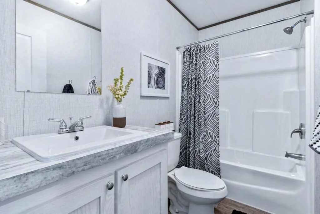 A clean, modern bathroom with a white sink, gray marble countertop, large mirror, small plant, framed art, and a toilet next to a bathtub with a black-and-white patterned shower curtain.