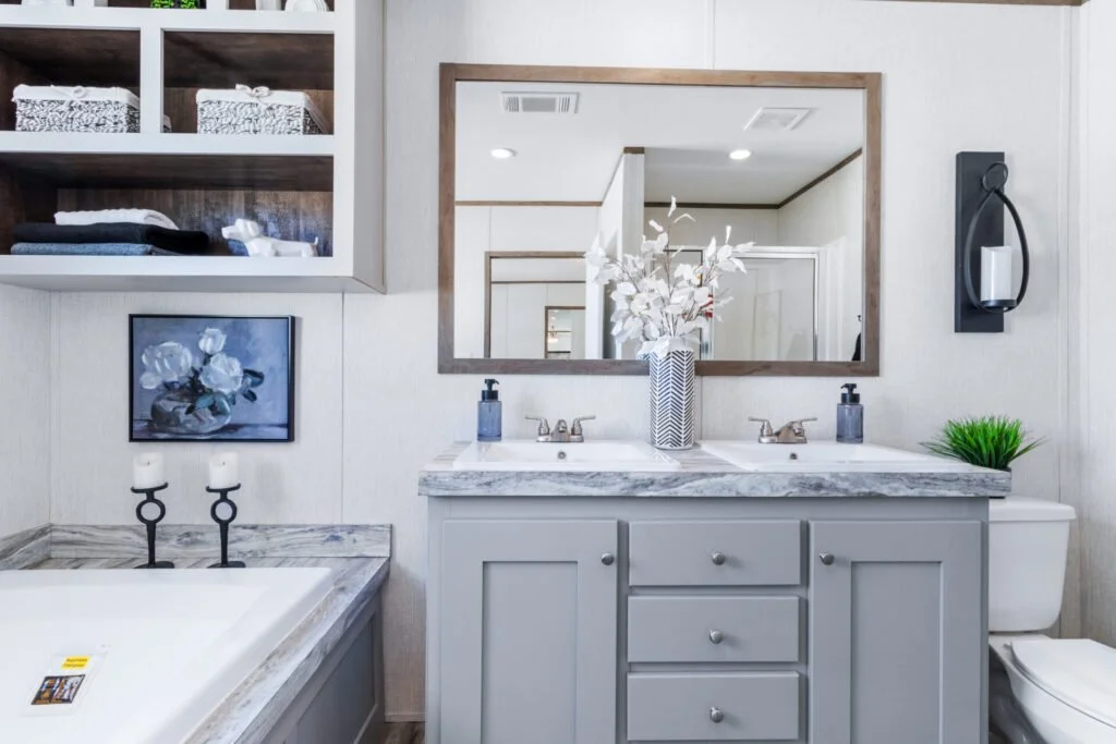 A modern bathroom with a double sink vanity, large mirror, and decorative items including a vase with white flowers, a small green plant, and a wall-mounted toilet paper holder with a roll of toilet paper. A framed floral painting and open shelves wi