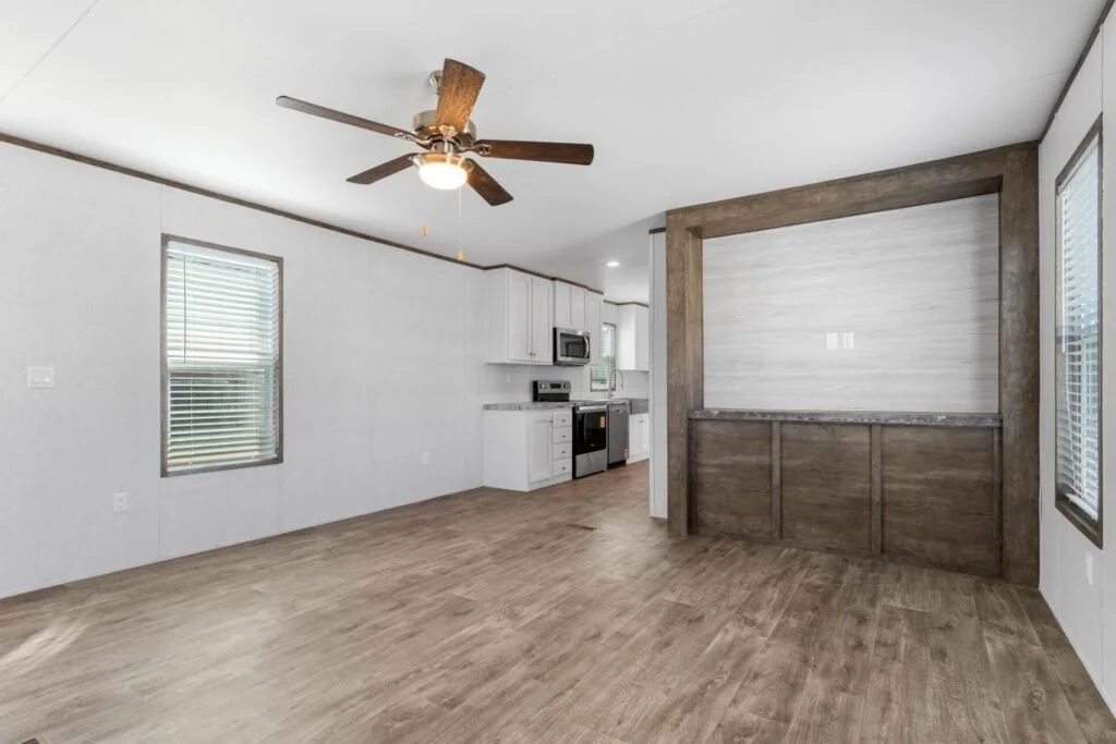 Empty living space with wood flooring, white walls, a ceiling fan, and a kitchen area with white cabinets and modern appliances.