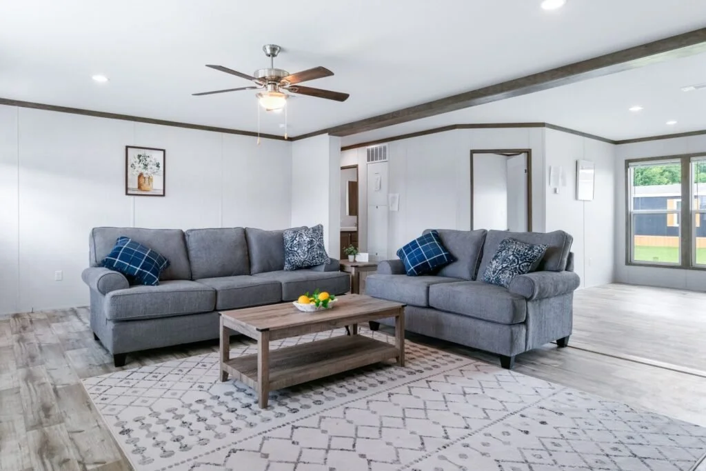Living room with two gray couches, blue patterned pillows, a wooden coffee table with a bowl of lemons, a ceiling fan, and large windows with a backyard view.
