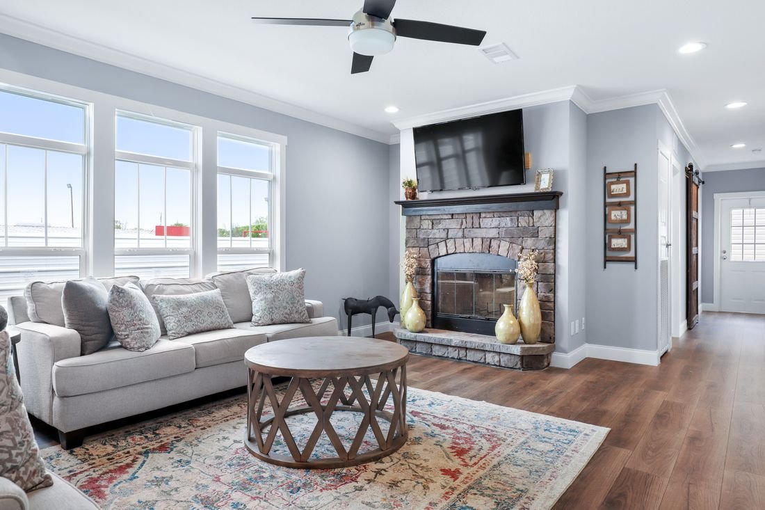 Bright living room with a gray sofa, wooden coffee table, stone fireplace with a mounted TV, potted plants, and large windows letting in natural light.