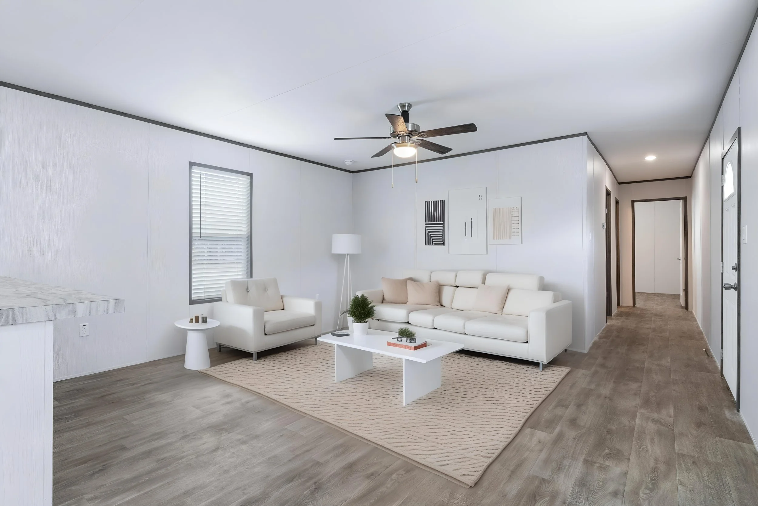 Living room with white sofa, beige chairs, coffee table, area rug, and artwork on white walls, with wood flooring and ceiling fan.