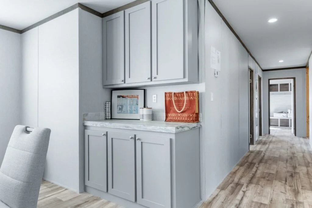 Interior view of a hallway with light gray walls, wood flooring, and white ceiling, leading to a room with a bed visible through the doorway.