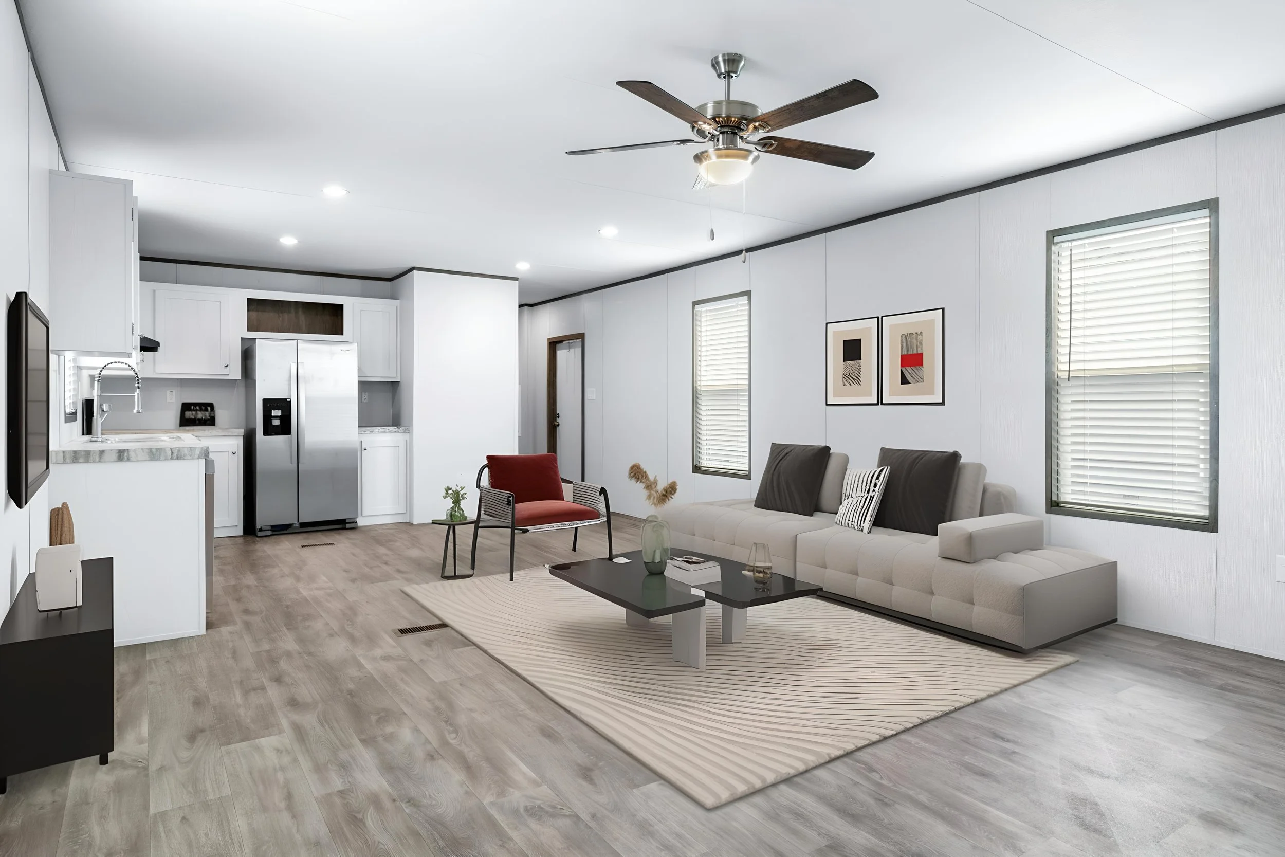 Living room with white walls, a beige sectional sofa, a black coffee table, and two framed abstract art pieces on the wall. There is a ceiling fan, windows with blinds, and a kitchen area in the background with white cabinets and a stainless steel re