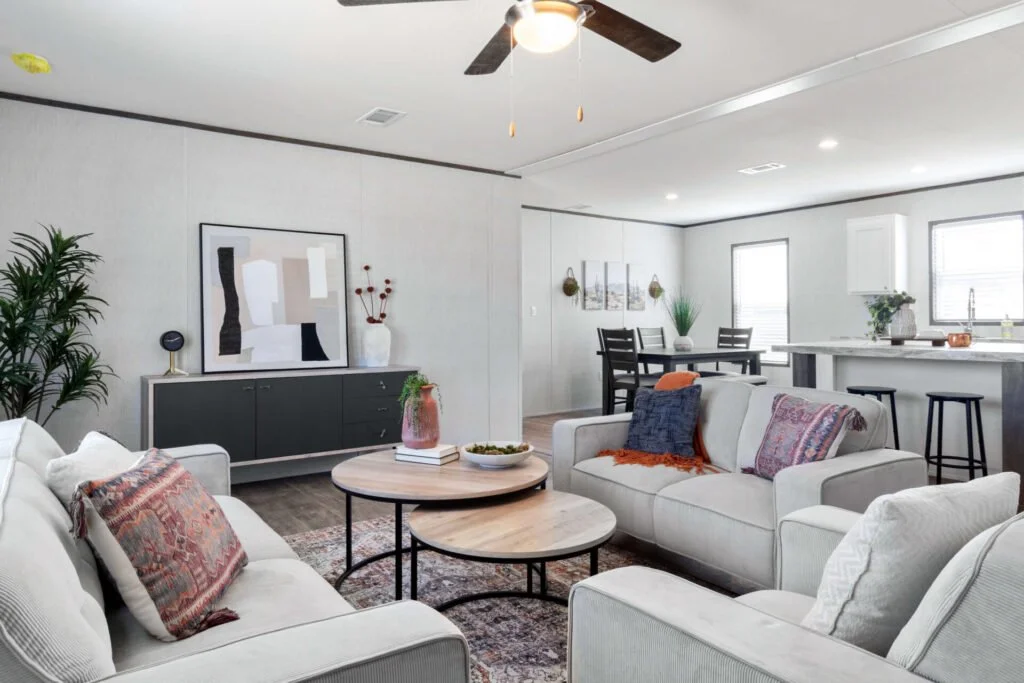 A modern living room with white walls, featuring sofas with decorative cushions, a round coffee table, a black console table with artwork and a vase, a dark dining table with chairs, large windows, and a kitchen with a white counter and bar stools.