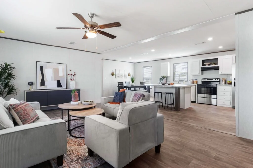 Open-concept living room and kitchen with white walls, hardwood floors, modern furniture, ceiling fan, and natural light from windows.