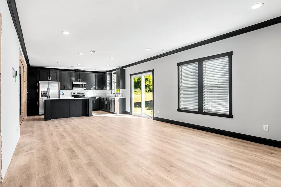 Empty living room with light wood flooring, black trim and window frames, white walls, black kitchen cabinets, stainless steel appliances, and a sliding glass door leading outside.