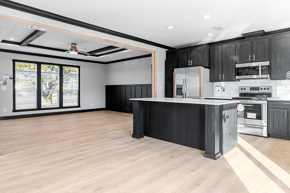 Open-concept living room and kitchen with black cabinetry, stainless steel appliances, light wood flooring, large windows, and a ceiling fan.