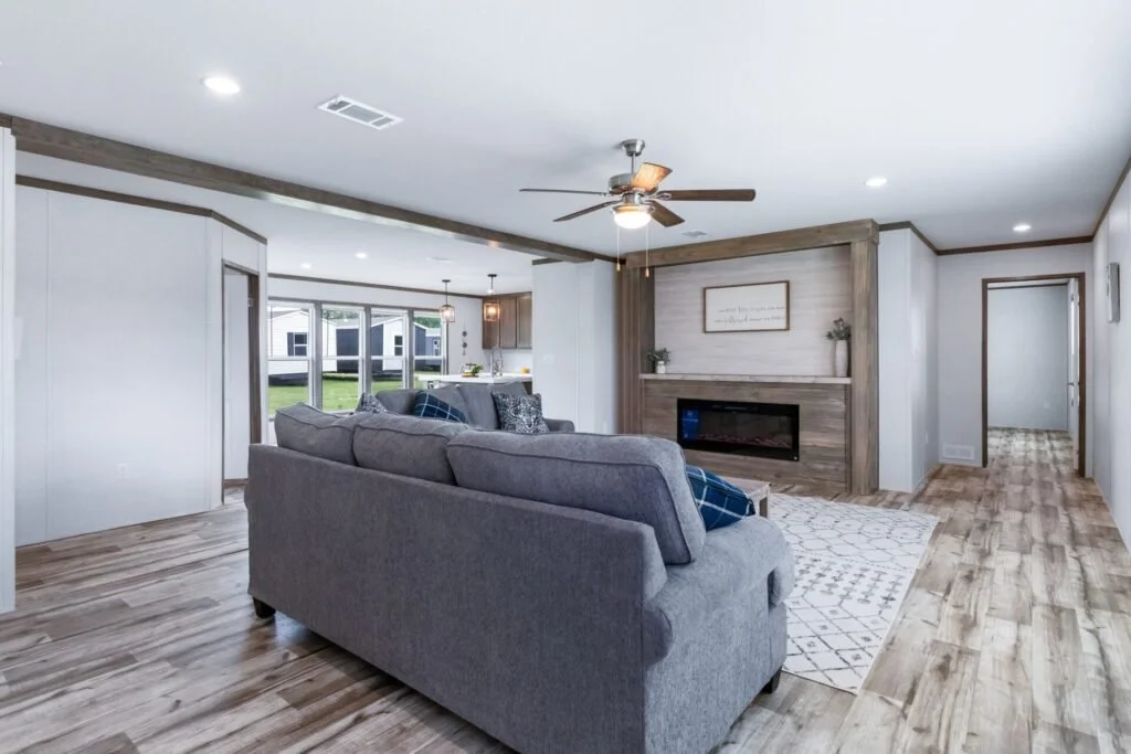 Modern living room with gray sofa, wood-burning fireplace, ceiling fan, hardwood flooring, and large windows.