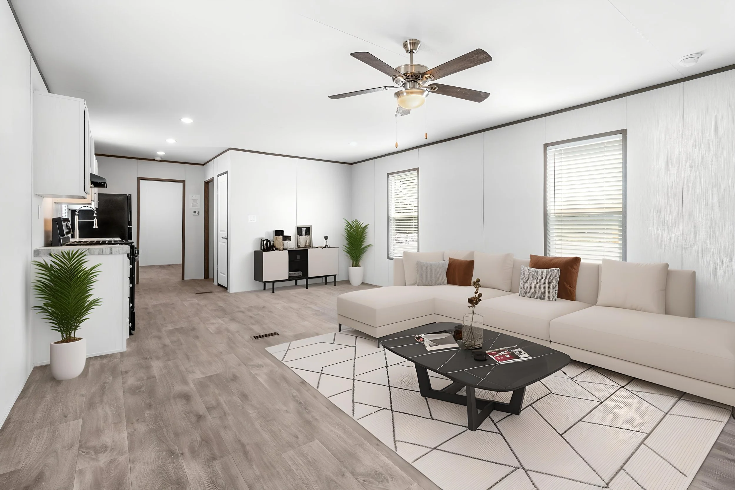 A spacious modern living room with a white sectional sofa, a black coffee table, and a geometric-patterned rug. The room has light wood flooring, white walls, two windows with blinds, and a ceiling fan with wooden blades. There is a small black and w