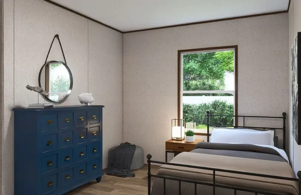 A bedroom with a metal bed frame, a wooden nightstand with a lamp and a plant, a blue dresser with a mirror and decor, a window showing green trees outside, and light-colored walls with a wooden floor.