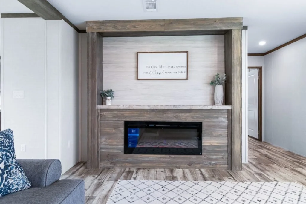 Living room with a modern electric fireplace built into a wooden feature wall, decorated with framed wall art and potted plants, with a gray sofa and a patterned rug in front.