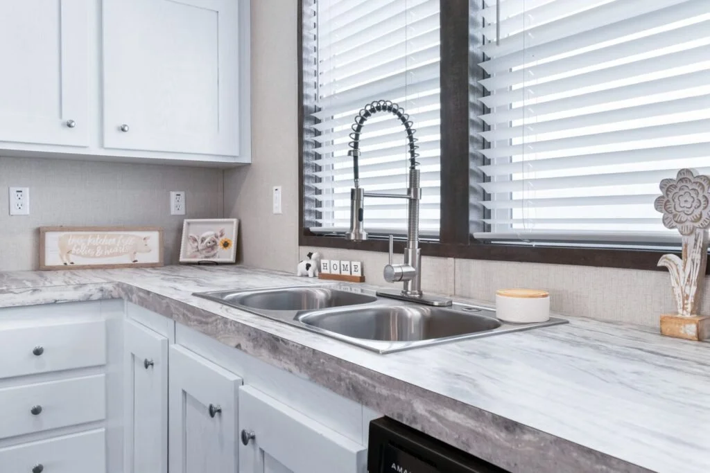 White kitchen countertop with a double stainless steel sink, a gooseneck faucet, and decorative items including a framed picture, a small wooden sign, a flower ornament, and a window with blinds.