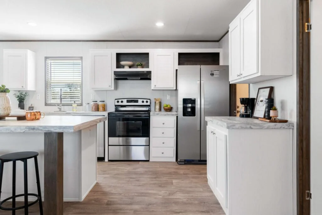 Modern kitchen with white cabinets, stainless steel appliances, and a wooden floor, featuring a small island with a black stool, and decorative items like potted plants and framed pictures.