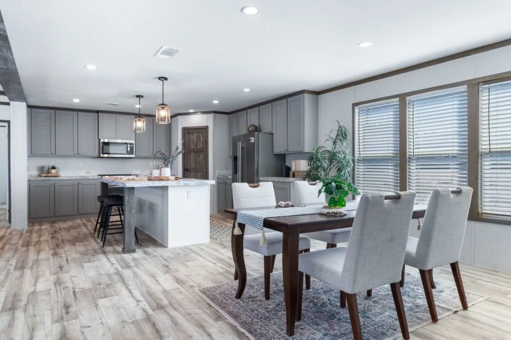 Modern kitchen with gray cabinets, stainless steel appliances, a kitchen island with bar stools, and a dining area with a wooden table, four upholstered chairs, and large windows with blinds.