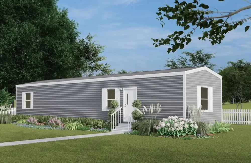 A single-story, gray manufactured home with white trim, surrounded by a landscaped lawn with flowers and bushes, and a white picket fence in the background.