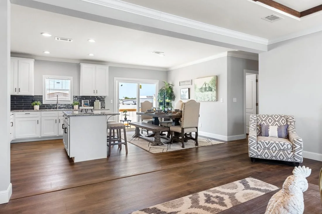 Open-concept kitchen and dining area with white cabinets, black backsplash, and hardwood floors, featuring a dining table with four beige chairs, a sliding glass door, and a patterned armchair.