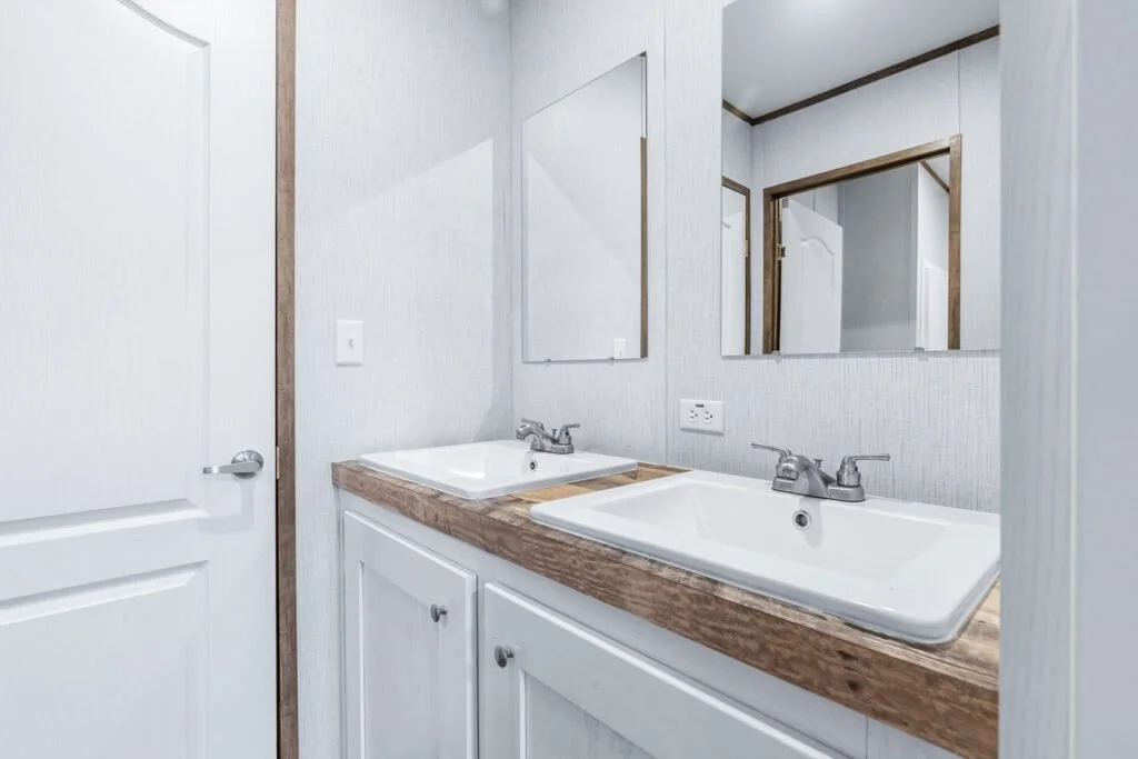 Bathroom with a double sink vanity, large mirror, white cabinets, light-colored walls, door, electrical outlet, and wood trim.