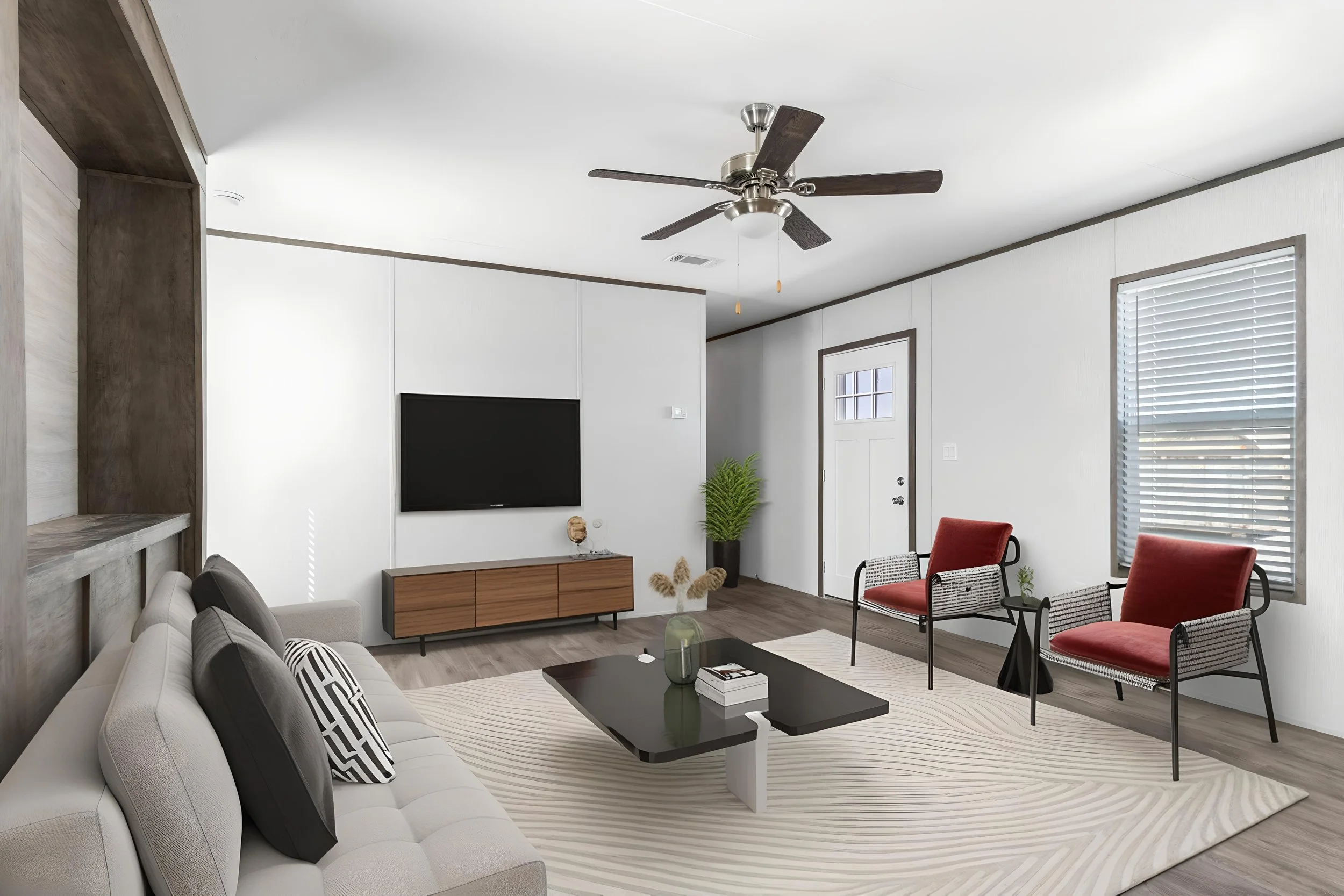 A modern living room with white walls, a ceiling fan, large windows with blinds, a flat-screen TV on a wooden stand, a white sofa with black and white pillows, a black coffee table with books and a vase, two red chairs, a plant in a black pot, and a 