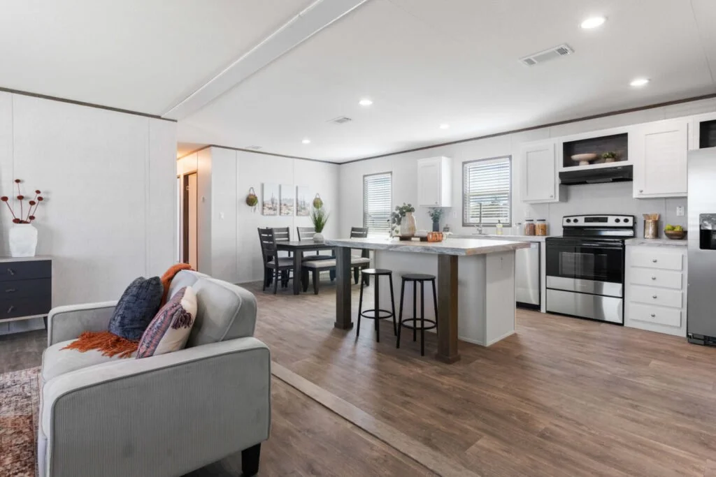 Open-concept living room and kitchen with white cabinets, stainless steel appliances, a kitchen island with two stools, a dining table with chairs, and decorative items including potted plants and artwork.