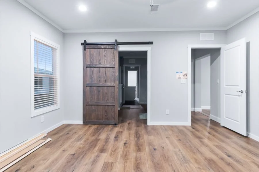 Empty room with wood flooring, white walls, a window with blinds, a wooden sliding barn door, and an open doorway leading to a hallway.