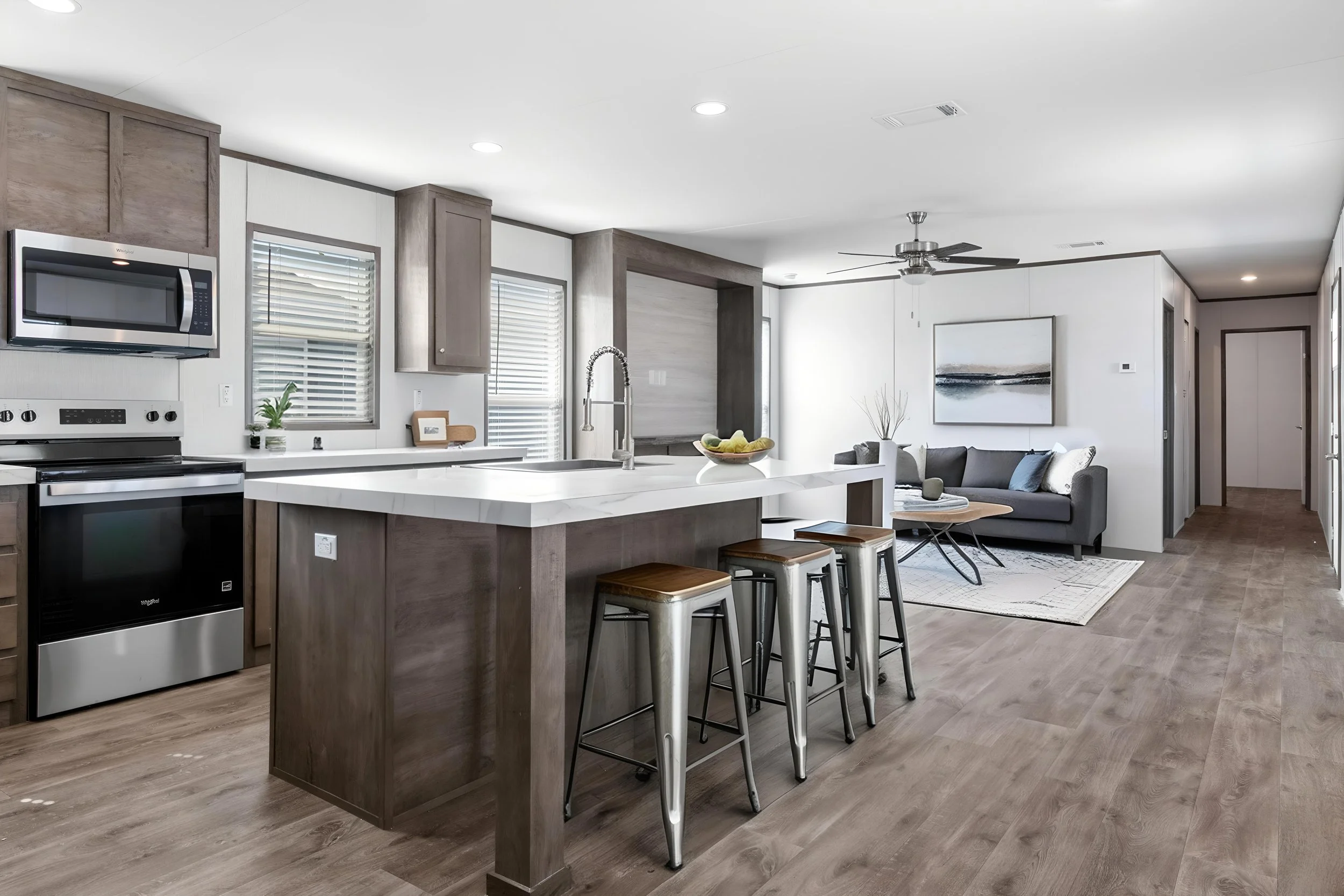 Open-concept kitchen and living room with wooden cabinetry, stainless steel appliances, a kitchen island with bar stools, and a sitting area with a gray couch and abstract wall art.