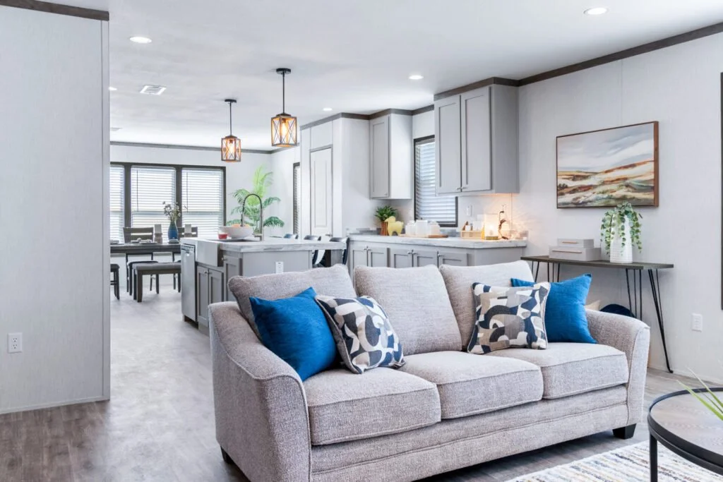 Living room with a beige couch, blue and patterned pillows, and a modern kitchen in the background with pendant lights and dining area.