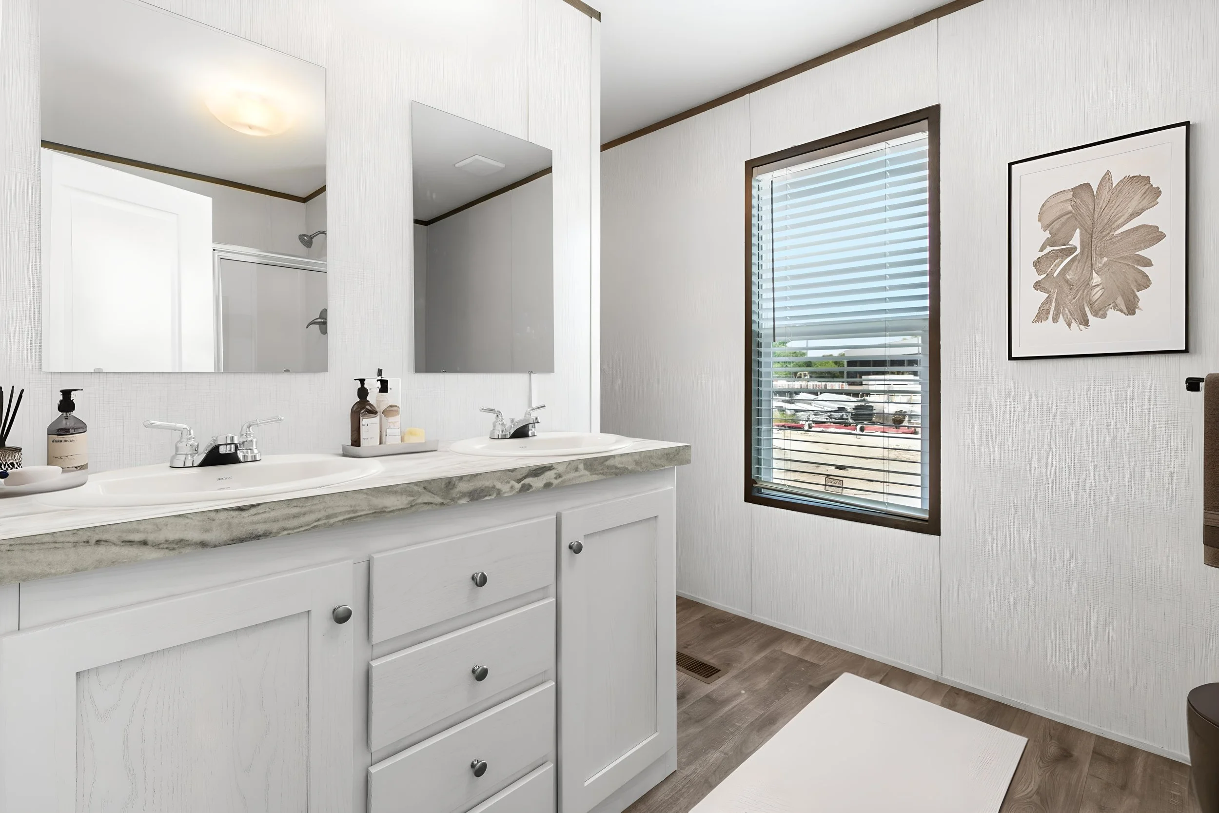 A bathroom with a double sink vanity with a marble countertop, two mirrors above the sinks, a window with blinds letting in natural light, and a framed floral artwork on the wall.