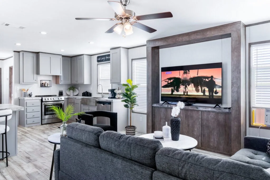 Modern living room with a TV, gray sofa, black and white furniture, kitchen with gray cabinets, and ceiling fan with lights.
