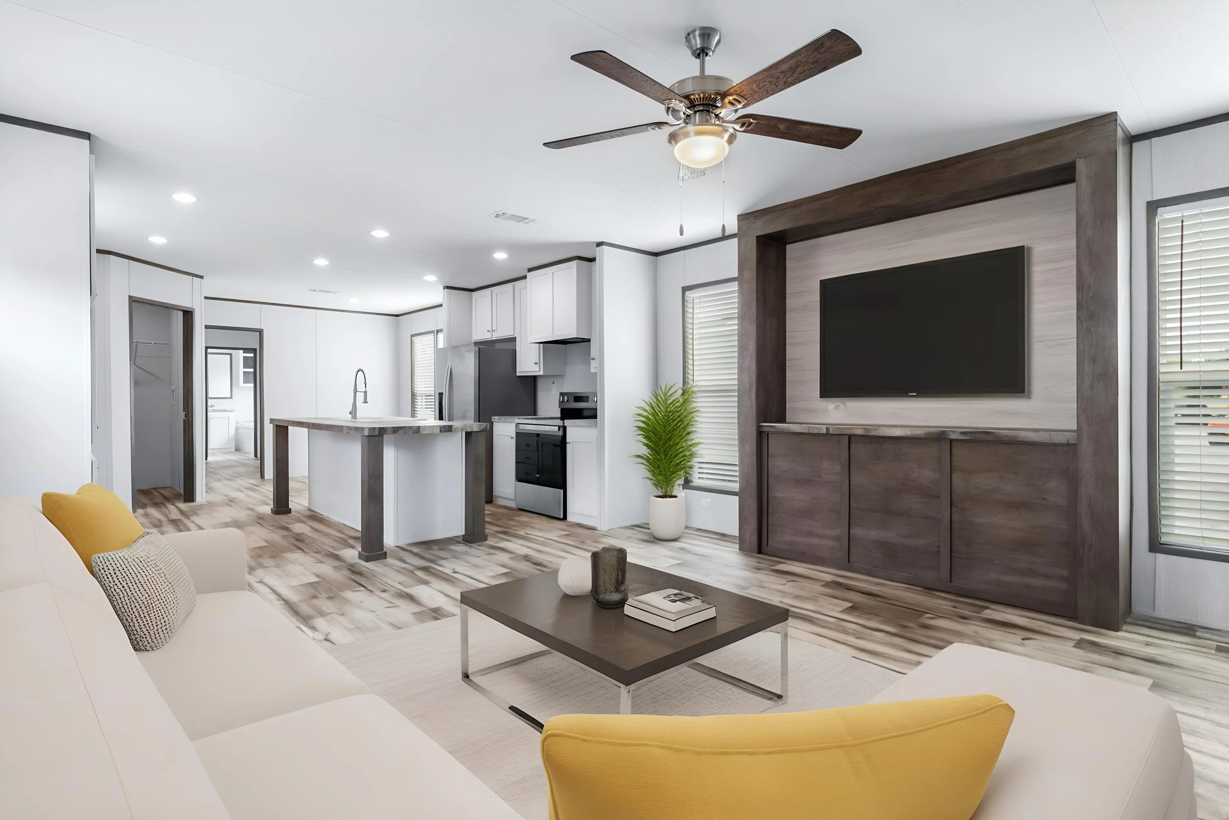 Modern open-concept living room and kitchen with white walls, wood flooring, a ceiling fan, and a large flat-screen TV mounted on a wooden accent wall. The space features a white sofa, yellow armchair, small coffee table, potted plant, and kitchen is