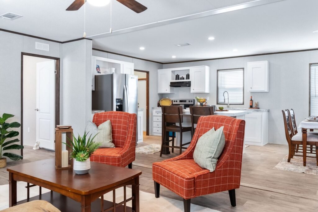 Open-concept living room with two orange plaid armchairs, a wooden coffee table, a kitchen with white cabinets, stainless steel refrigerator, dining table, and natural light from windows.