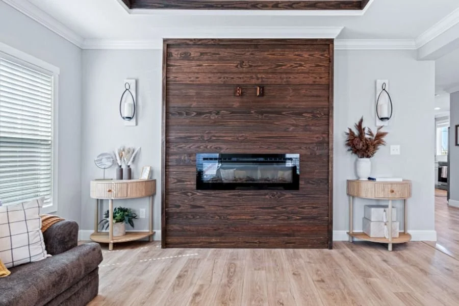 Living room with a built-in fireplace on a brown wood accent wall, two round side tables with decor, a brown sofa with pillows, and a window with white blinds.