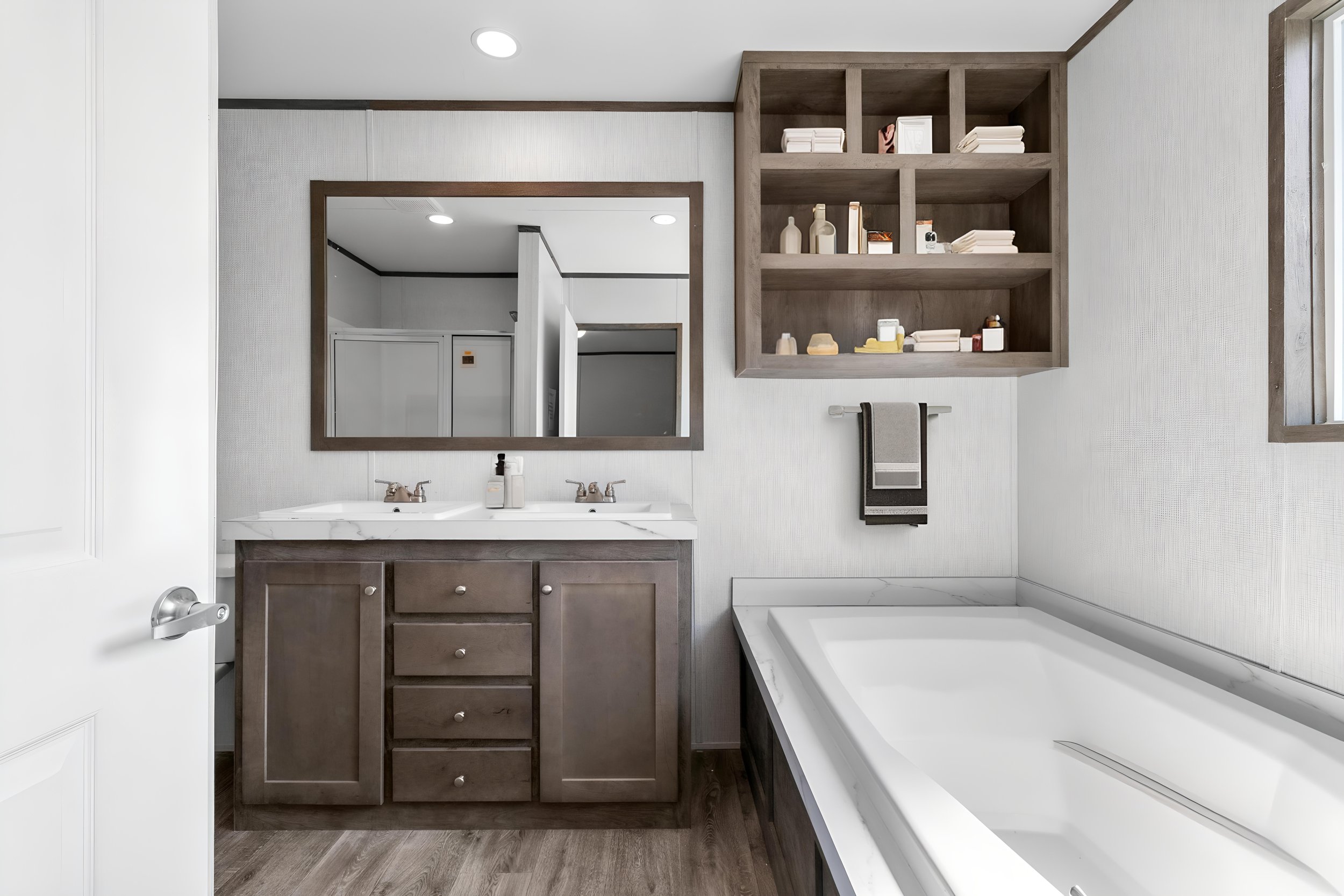 A bathroom with a white vanity, two sinks, and a large mirror above it. There are open shelves with toiletries and towels. A bathtub is visible on the right side.