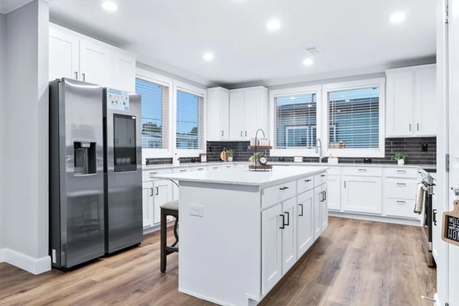 Modern kitchen with white cabinets, stainless steel refrigerator, large island, black backsplash, and hardwood floors.