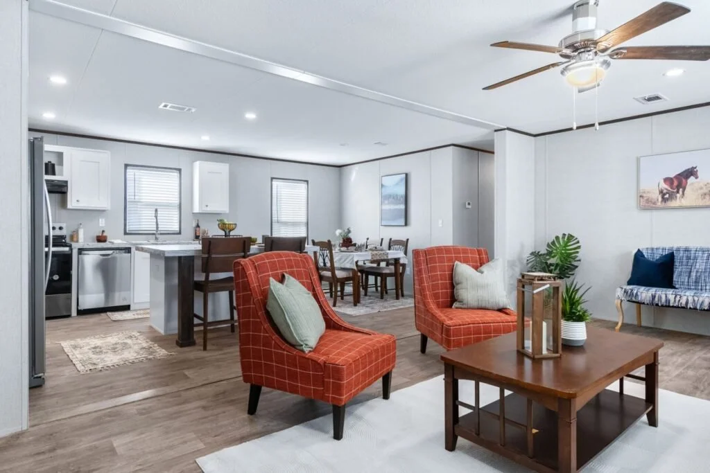 Open-concept living room and kitchen with white walls, hardwood floors, two red armchairs with pillows, a wooden coffee table with decorative items, a bench with throw pillows, and a ceiling fan.