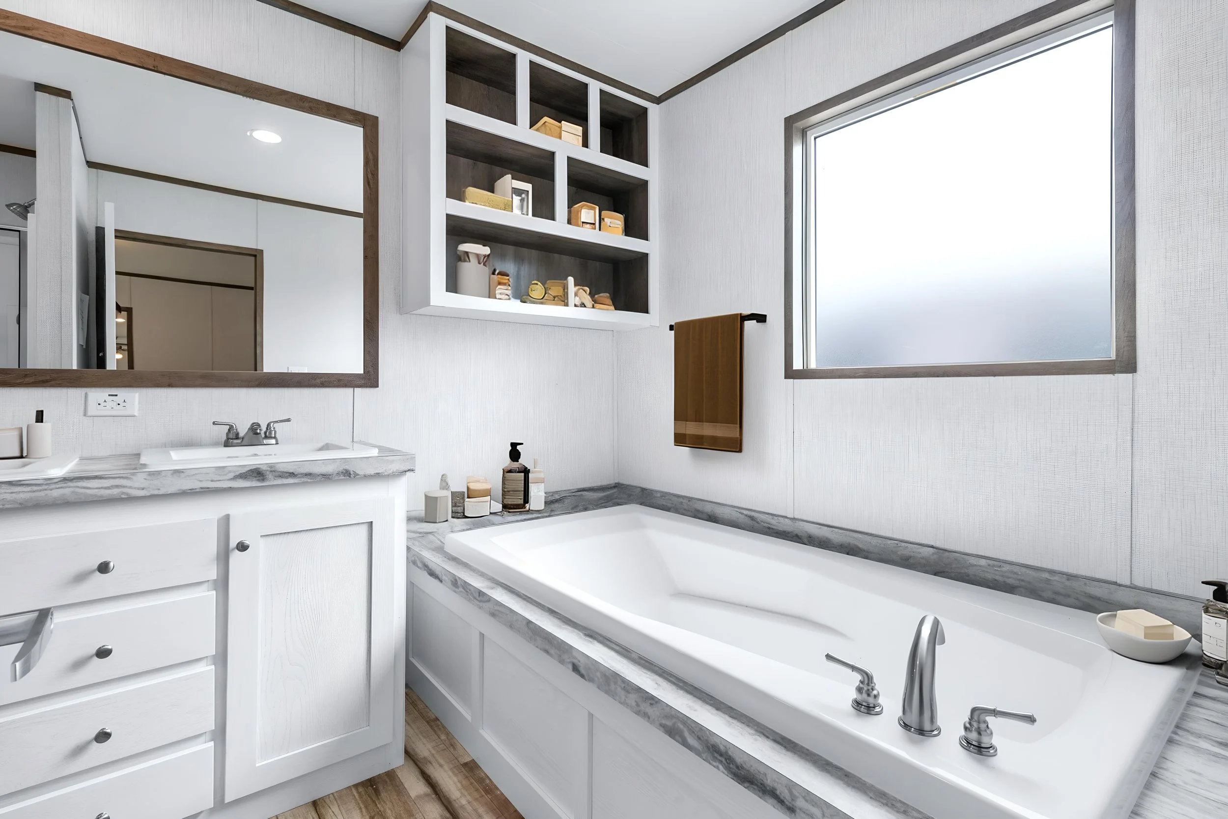 A modern bathroom with a large frosted window, white cabinetry, a marble countertop with a sink, and decorative bottles. There is a mirror on the wall, a towel hanging on a black towel rod, and a built-in shelf with toiletries and decorative items.
