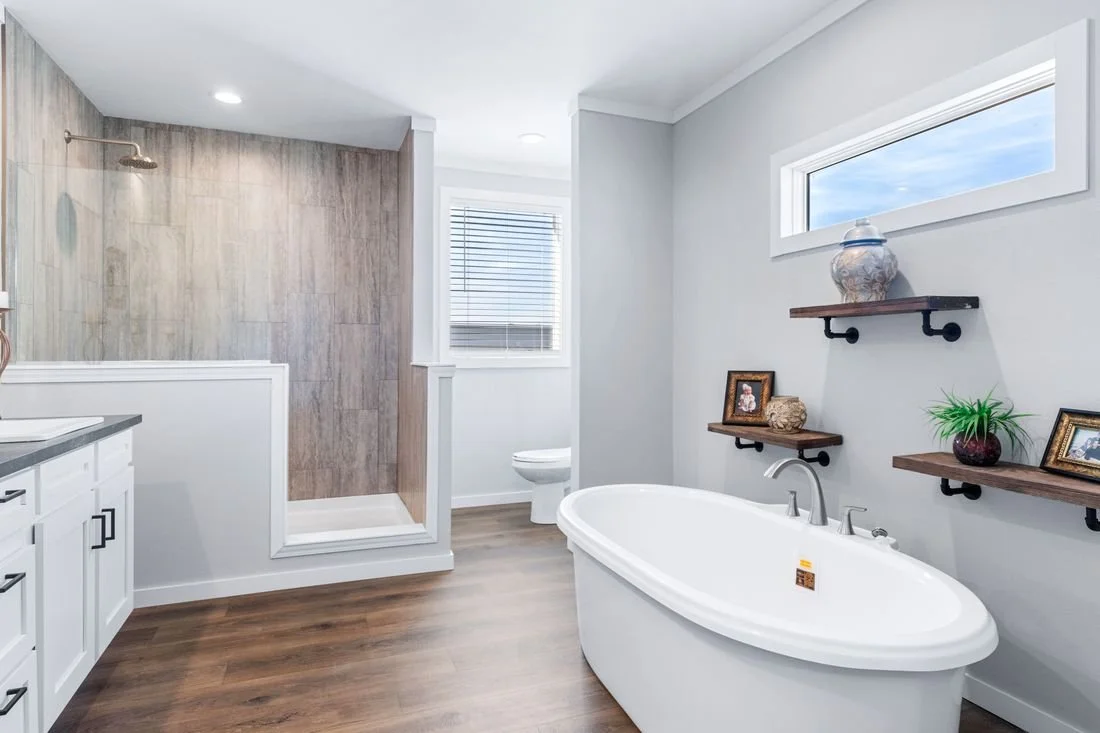 Modern bathroom with a walk-in shower, a white freestanding bathtub, and a toilet. The room has light gray walls, wooden floating shelves with decorative items, and wood-look flooring. There is a window above the bathtub and another window near the s