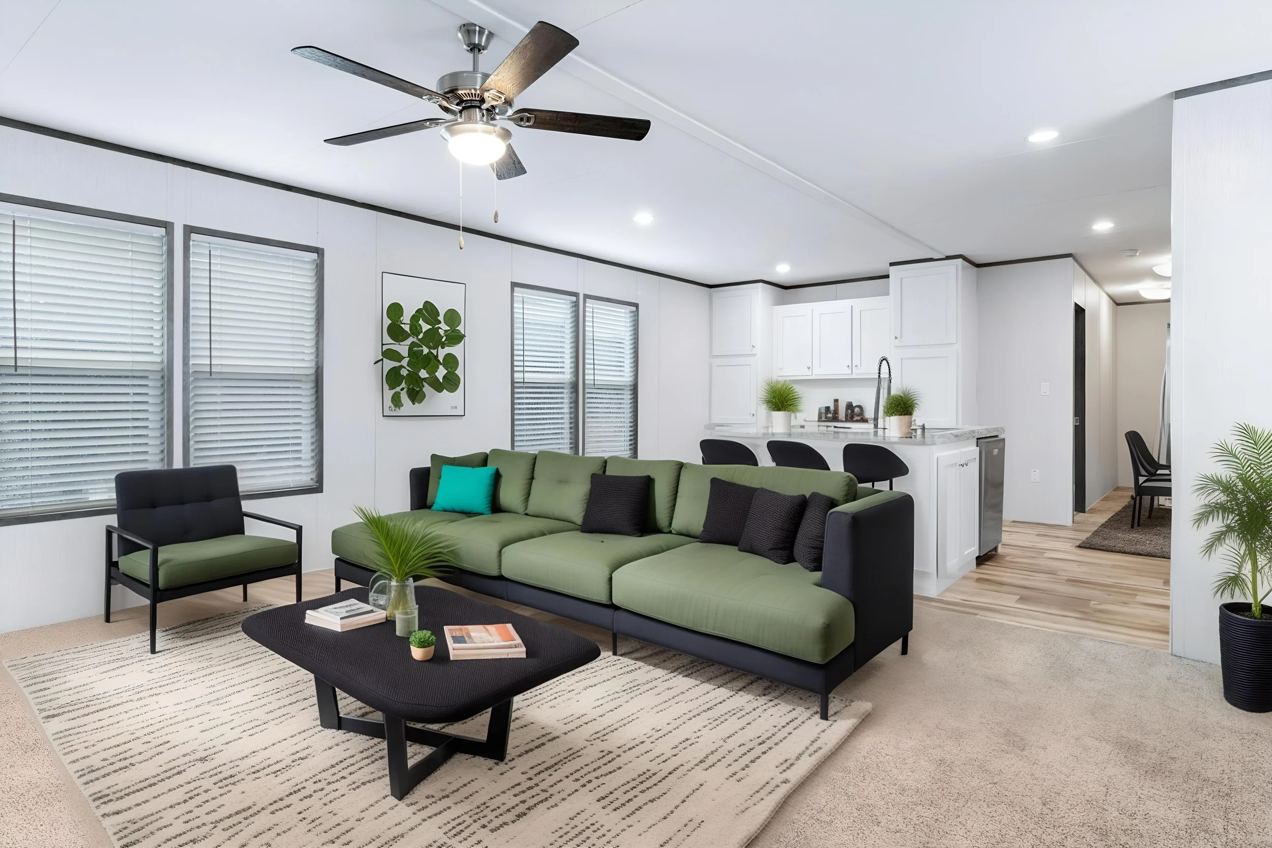 Living room with white walls, three large windows with blinds, a green sectional sofa, black armchair, patterned area rug, modern coffee table with books and a small plant, in an open-plan space with a kitchen in the background, ceiling fan, pendant 
