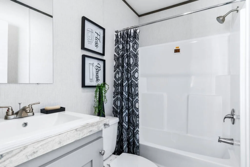 A bathroom with a white bathtub and shower, a small white vanity with a silver faucet, two framed art prints on the wall, a green potted plant, and patterned curtains, with a mirror closed on the left.