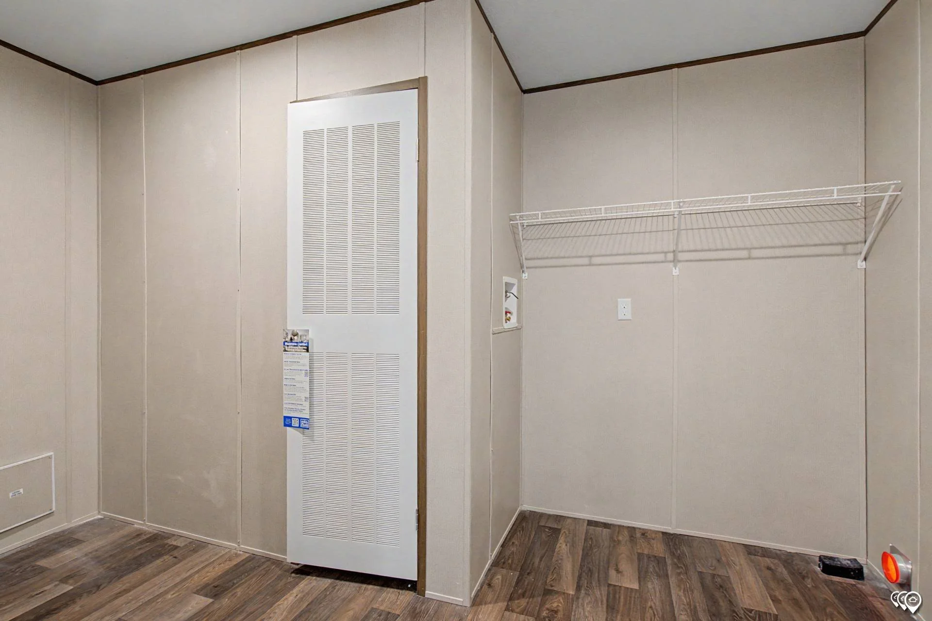Empty room with beige wall panels, a white wire shelf mounted on the wall, a white vent cover on a door, and a wood-tone floor.